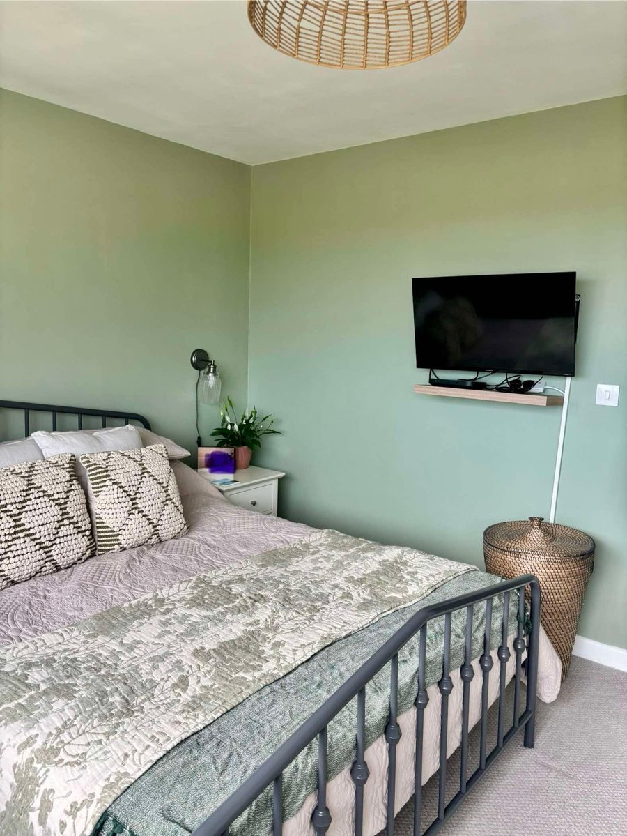 Bedroom painted in sage green with a wall mounted television and a floating shelf fitted beneath it.